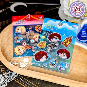 Peut inclure: Deux planches d'autocollants "PUPPET PALS" avec des illustrations de personnages de style anime et des objets thématiques. L'une a un fond rouge, l'autre bleu. L'image comprend également un autocollant "HUMAN MADE ART" et le texte "Cosmic Whale Cafe".