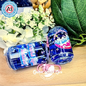 May include: Two blue, cylindrical tumblers with the text "Trans Live Matter" and "Transpo Cream" in a playful font. The tumblers are decorated with stars and a pink keychain. The background includes white flowers and green leaves.