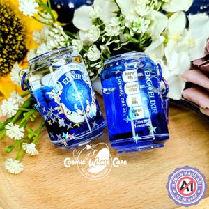Puede incluir: Dos pequeños frascos de vidrio transparente llenos de líquido azul. Uno tiene el texto "BO ELIXIR" y un diseño azul con estrellas. El otro tiene el texto "ERGO ELIXIR" e información nutricional. Ambos tienen asas de metal plateado.