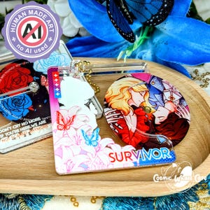 May include: A collection of clear acrylic keychains and charms with various designs. One keychain features the word "SURVIVOR" and a floral and butterfly design. Other charms have illustrations of people and roses. The keychains are on a wooden tray.
