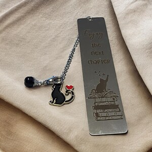 May include: A silver metal bookmark with a black cat charm and the words "Enjoy the next chapter." The bookmark features a black cat illustration on a stack of books. A black bead and a red heart charm are also present.