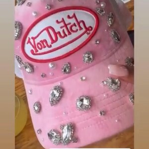 May include: A pink Von Dutch trucker hat, adorned with rhinestones and pearls. The hat has a white mesh back and a pink front panel with the Von Dutch logo. A fashionable accessory.