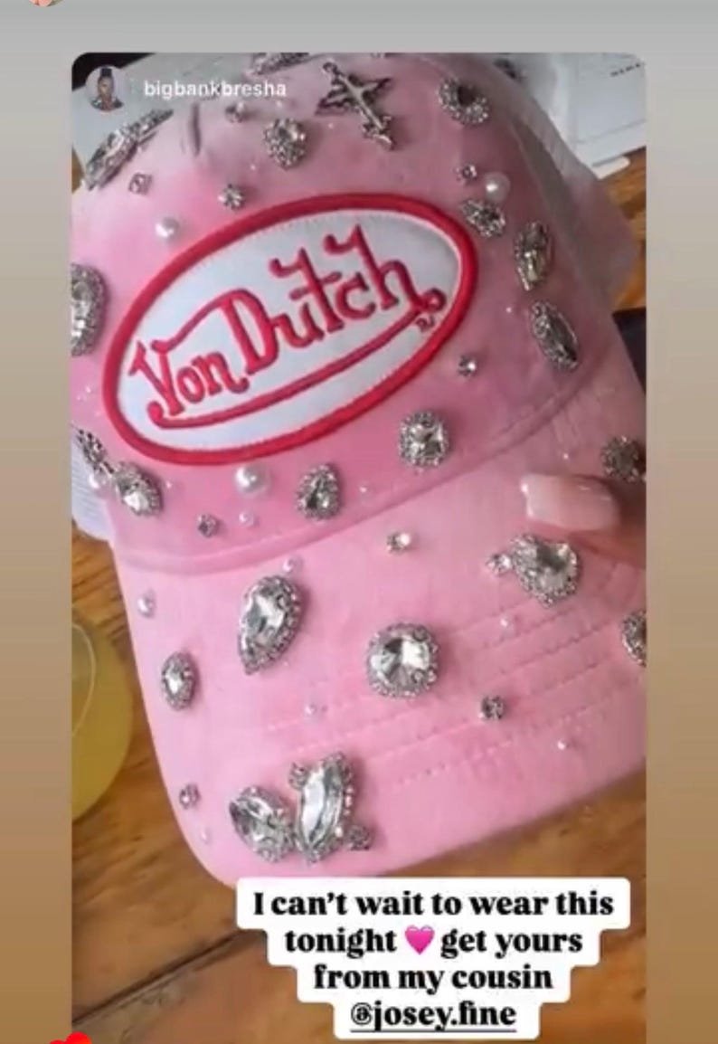 May include: A pink Von Dutch trucker hat, adorned with rhinestones and pearls. The hat has a white mesh back and a pink front panel with the Von Dutch logo. A fashionable accessory.