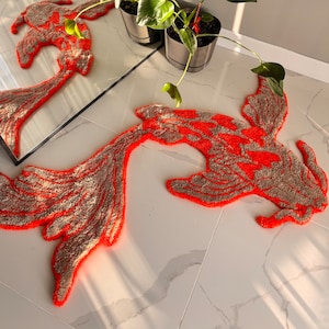 May include: A tufted rug shaped like a koi fish, featuring a vibrant neon orange outline and intricate beige textured patterns throughout its body and flowing tail. The rug is laid flat, showcasing its detailed organic form and soft fibre construction.