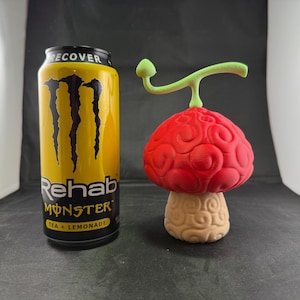 May include: A red and white 3D printed mushroom with a green stem. The mushroom is about 5 inches tall and is sitting on a black surface. The mushroom is a replica of the "Hito Hito no Mi" Devil Fruit from the anime series "One Piece".