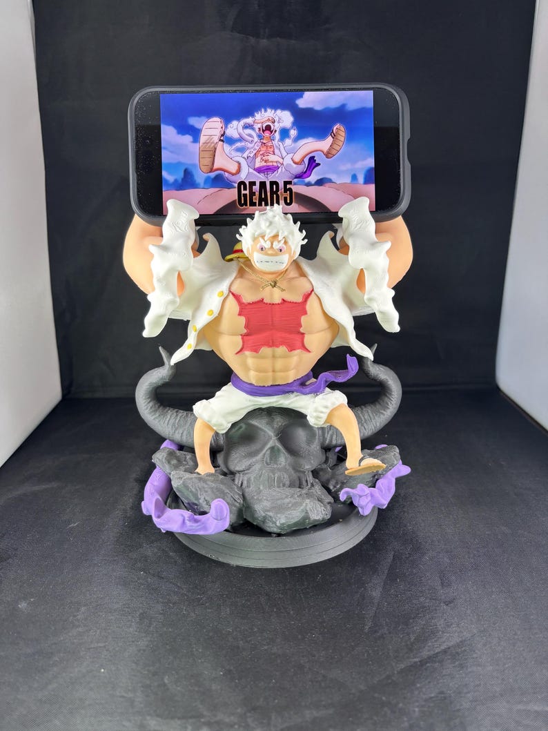 May include: A 3D printed phone stand featuring a character from the anime One Piece. The character is in a white and red outfit with a skull and purple flames at the base. The phone stand has the text "GEAR 5" on it.