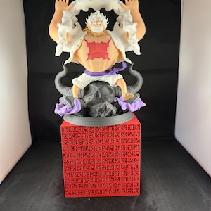 May include: A white and red anime figurine of a character with white hair and a white shirt. The character is standing on a black and purple base with a skull design. The base is on a red cube with black text.
