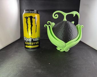 May include: A green and black 3D printed model of a fruit with a black sphere surrounded by green leaves. The model is sitting on a black surface next to a can of Monster Rehab Tea + Lemonade.