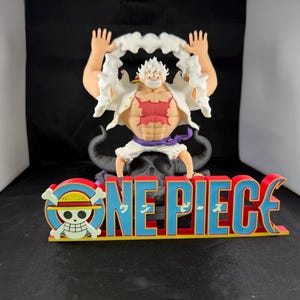 May include: A white and red figure with white hair and a white shirt with a red design is sitting on a black figure with horns. The figure is holding a white cloud in each hand. The figure is sitting on a red and blue "ONE PIECE" sign with a skull and crossbones logo.