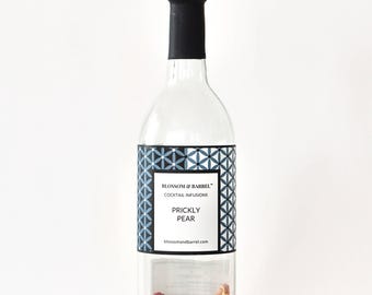 May include: A clear glass bottle with a black cap and a label that reads "Blossom & Barrel" and "Prickly Pear." The bottle contains dried fruit and spices. The label has a geometric design in blue and white.