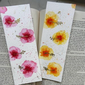 May include: Two watercolor bookmarks with floral designs. The bookmarks are painted on white paper with pink and yellow flowers. The flowers have gold accents and are surrounded by gold splatters.