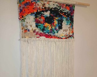 May include: A colorful woven wall hanging with a geometric design and white fringe. The hanging is made with yarn and is attached to a wooden dowel.