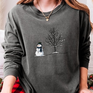 May include: Dark gray long-sleeve shirt featuring a snowman with a black hat and blue scarf next to a bare tree. The snowman has a carrot nose and button eyes. The shirt is made of a soft material.