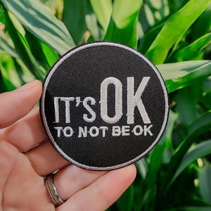 Può includere: Una toppa rotonda nera con testo ricamato bianco che recita "IT'S OK TO NOT BE OK". La toppa ha un bordo bianco. L'immagine mostra la toppa su uno sfondo di fogliame verde.