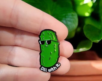 May include: A green enamel pin in the shape of a pickle wearing black sunglasses. The pin has a black border and the words "Dill with it" in white text.