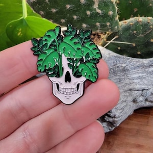 May include: A white skull-shaped enamel pin with a black outline and a green plant design. The pin features a skull with green leaves and stems growing from the eye sockets and top of the skull. The pin is held in a hand.