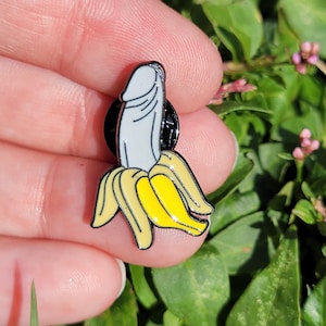 May include: A novelty enamel pin depicting a banana with a phallic design. The pin is white and yellow with black detailing. The pin is set against a backdrop of green leaves and foliage.