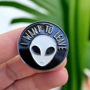 Pode incluir: Um broche de esmalte preto e branco com um rosto de alienígena branco e o texto "I want to leave" em um círculo ao redor do alienígena.