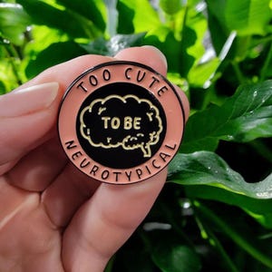 May include: A circular enamel pin with a peach-coloured border and black centre. The pin reads "TOO CUTE TO BE NEUROTYPICAL" in white text. A gold-coloured brain graphic is in the centre of the pin.