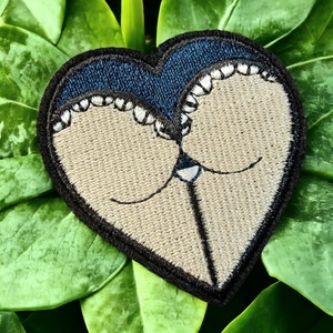 Pode incluir: Um remendo bordado em forma de coração com um design azul e branco. O remendo é feito de feltro e tem um contorno preto.
