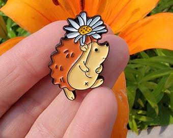 Puede incluir: Un pin de esmalte caprichoso con un erizo de dibujos animados sosteniendo una margarita. El pin tiene un cuerpo amarillo, una espalda naranja y una margarita blanca y amarilla. El pin se sostiene en una mano contra un fondo de flores naranjas.