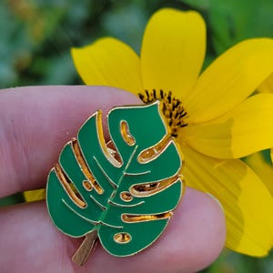 May include: A green and gold enamel pin in the shape of a monstera leaf. The pin has a gold outline and a bright green interior. The pin is held in front of a yellow flower.