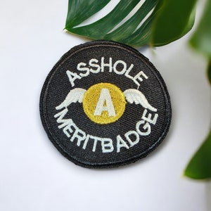May include: Black round patch with white embroidered text that reads "Asshole Merit Badge" and a gold letter "A" with white wings.