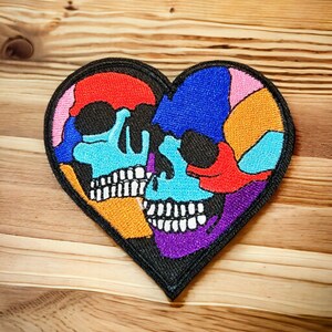 Può includere: Un cerotto ricamato colorato a forma di cuore con due teschi all'interno. I teschi sono fatti di pezzi di tessuto di diversi colori, tra cui rosso, blu, arancione, giallo, viola e bianco.