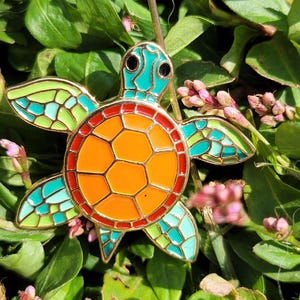 Puede incluir: Un colorido broche de esmalte con forma de tortuga con un contorno dorado. La tortuga presenta un caparazón naranja brillante, cabeza y aletas turquesas y detalles verdes. El broche se encuentra sobre un fondo de hojas verdes y pequeñas flores rosas.