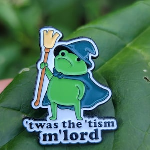 Puede incluir: Un pin de esmalte de una rana verde con un sombrero de mago y una capa de color azul verdoso oscuro, sosteniendo una escoba color canela. Debajo de la rana, una cinta blanca con borde negro muestra el texto negro 'twas the 'tism m'lord'.
