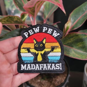 Op de afbeelding: Geborduurde patch met een zwart kattenontwerp, met pistolen en de woorden "PEW PEW MADAFAKAS!". De patch heeft een achtergrond in zonsondergangstijl met rode, oranje, gele en blauwe strepen. De patch is zwart met een witte rand.