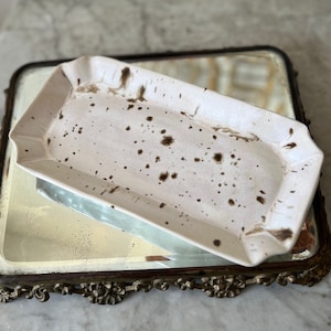 May include: A white ceramic rectangular tray with brown speckles sits on a gold-colored decorative tray with a mirrored surface.