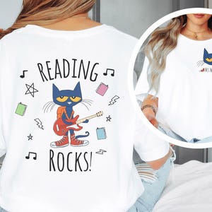 May include: White t-shirt with a blue cat playing a guitar and the text "Reading Rocks!" printed on the back. The cat is wearing a backpack and red shoes. There are musical notes, stars, and books surrounding the cat.