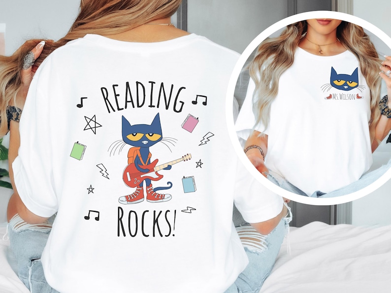 May include: White t-shirt with a blue cat playing a guitar and the text "Reading Rocks!" printed on the back. The cat is wearing a backpack and red shoes. There are musical notes, stars, and books surrounding the cat.
