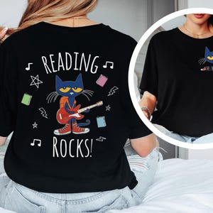 May include: Black t-shirt with a graphic of a blue cat playing a red guitar. The text "Reading Rocks!" is printed on the shirt. The cat is wearing a backpack and has a yellow star on its forehead. The shirt has a musical note and lightning bolt designs.
