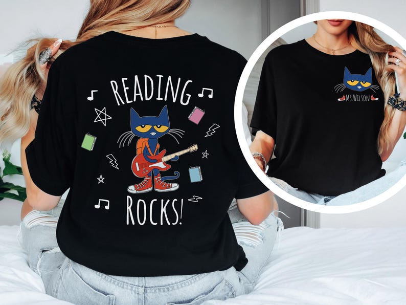 May include: Black t-shirt with a graphic of a blue cat playing a red guitar. The text "Reading Rocks!" is printed on the shirt. The cat is wearing a backpack and has a yellow star on its forehead. The shirt has a musical note and lightning bolt designs.