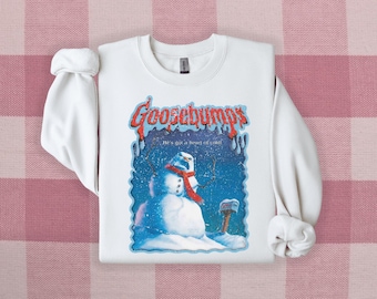 Puede incluir: Sudadera blanca con un gráfico de portada de libro de Goosebumps que presenta un muñeco de nieve con una bufanda roja y el texto "¡Tiene un corazón de frío!"