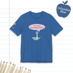 May include: A blue short-sleeve t-shirt featuring a cartoon pigeon graphic with a pink speech bubble containing the text Oh, hello kindergarteners. The sleeve displays a small icon of a magnifying glass and the text Smith in white lettering.