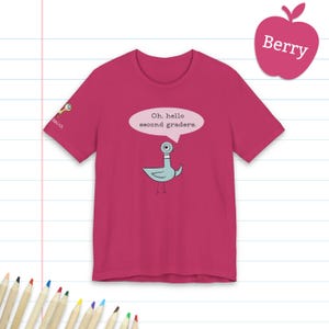 May include: A berry-colored short-sleeve t-shirt featuring a graphic of a light blue pigeon with a speech bubble that says, Oh, hello second graders. The sleeve includes a small magnifying glass icon and the text Smith.