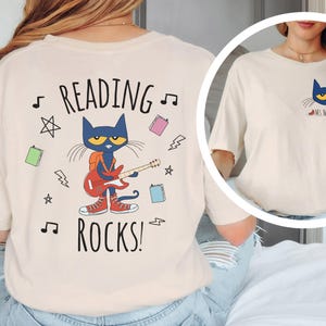 May include: A beige t-shirt with a graphic of a blue cat playing a guitar and the text "Reading Rocks!" on the back. The front of the shirt has a smaller graphic of the blue cat with the text "Ms. Wilson".