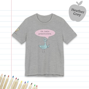 May include: A heather grey short-sleeve t-shirt featuring a light blue cartoon pigeon with a single large eye. Above the pigeon is a pink speech bubble containing the black text, Oh, hello preschoolers. A small key icon is on the sleeve.
