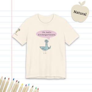 May include: A natural-colored t-shirt featuring a blue cartoon pigeon illustration with a pink speech bubble containing the text "Oh, hello kindergarteners." A small magnifying glass icon and the word "Smith" appear on the right sleeve.