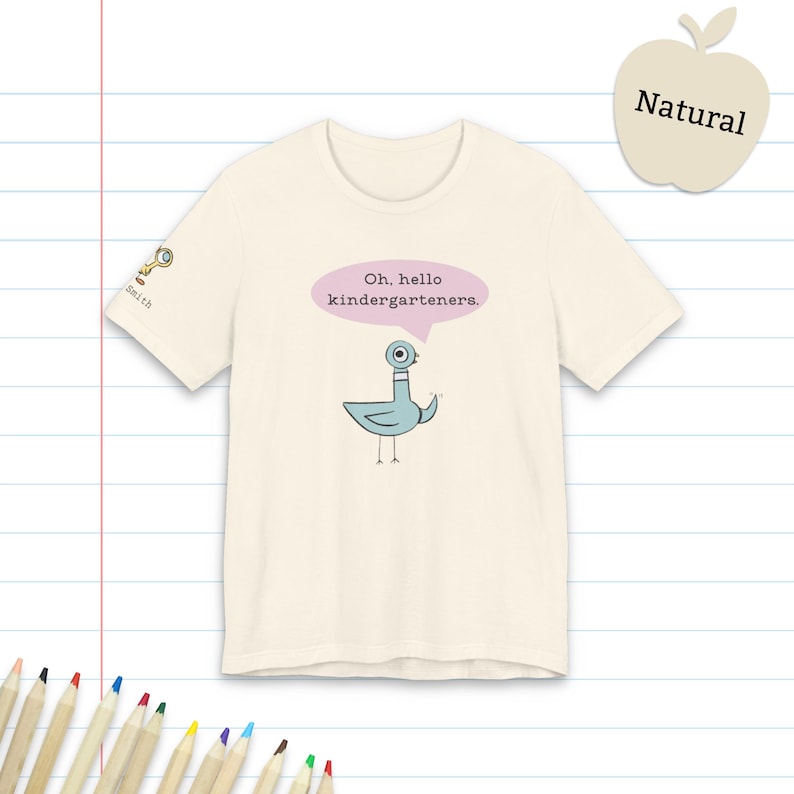 May include: A natural-colored t-shirt featuring a blue cartoon pigeon illustration with a pink speech bubble containing the text "Oh, hello kindergarteners." A small magnifying glass icon and the word "Smith" appear on the right sleeve.