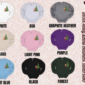 May include: A selection of Gildan 18000 unisex heavy blend crewneck sweatshirts in various colors, including white, ash, graphite heather, sand, light pink, purple, light blue, black, and forest. Each sweatshirt features a small graphic of a Christmas tree with a person standing next to it.