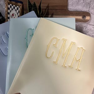 May include: Three folders in pastel shades of blue, mint green, and cream. Each folder features a large, decorative embroidered monogram on the front cover. The cream folder displays the letters CMA, while the others show partial monograms.
