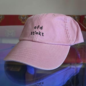 Puede incluir: Una gorra de béisbol de color rosa claro con el texto "afd stinkt" bordado en negro en el panel frontal. La gorra cuenta con una visera curva con costuras visibles y una corona estructurada, presentando un diseño sencillo y limpio.