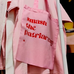 Puede incluir: Una bolsa de tela de algod&oacute;n rosa claro con el texto rojo "smash the patriarchy" impreso en la parte delantera, colgada sobre una camiseta de manga corta rosa claro a juego. La bolsa tiene dos asas de tela y un peque&ntilde;o punto rojo decorativo.