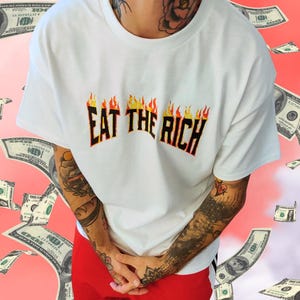 Puede incluir: Camiseta blanca de cuello redondo con el texto "EAT THE RICH" en un diseño de llama. La camiseta se combina con pantalones rojos. El fondo presenta billetes de dólar cayendo sobre un fondo rosa.