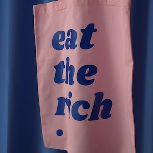 Puede incluir: Una bolsa de tela de color rosa claro con el texto "eat the rich" impreso en una fuente azul oscuro llamativa. La bolsa tiene dos asas de tela rosa a juego en la parte superior y un peque&ntilde;o punto azul oscuro debajo del texto a la izquierda.