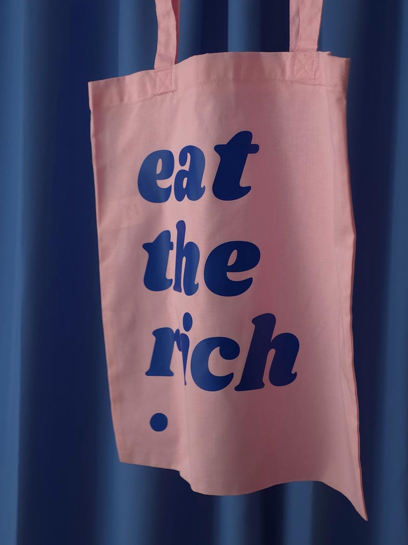Puede incluir: Una bolsa de tela de color rosa claro con el texto "eat the rich" impreso en una fuente azul oscuro llamativa. La bolsa tiene dos asas de tela rosa a juego en la parte superior y un peque&ntilde;o punto azul oscuro debajo del texto a la izquierda.